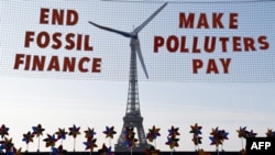 Replika turbin angin terpasang di sebuah jaring yang bertulisan "Hentikan pendanaan energi fosil, pencemar harus membayar" di tengah demonstrasi memprotes pendanaan energi fosil di Paris, 21 Juni 2023. (Foto: Ludovic Marin/AFP)