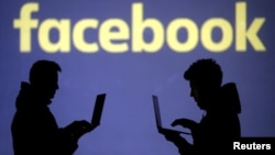 Silhouettes of laptop users are seen next to a screen projection of Facebook logo in this picture illustration taken March 28, 2018. 